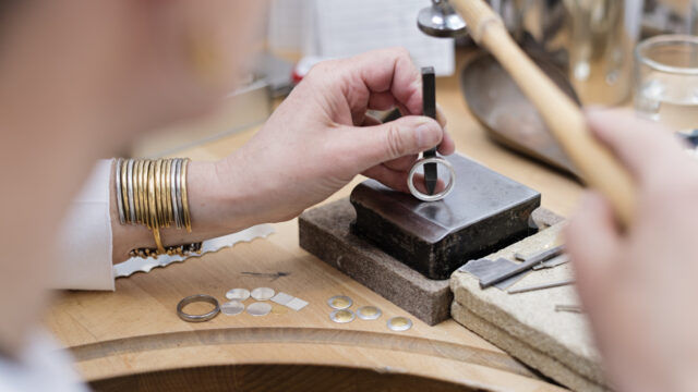 Portrait of a professional Goldsmith at work in her workshop creating a silver ring. In this picture she is stamping her hallmark on the inside on the ring. She is surrounded by the tools she uses to create her pieces of jewellery.