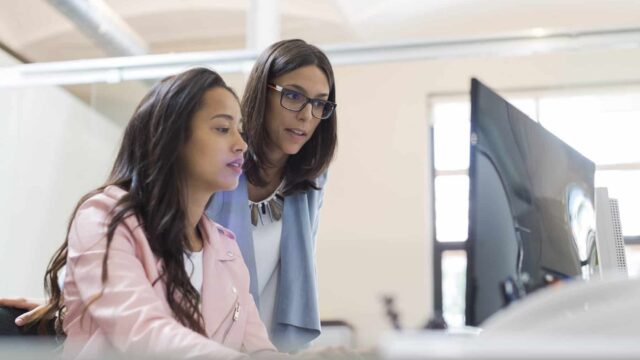 Two women look at a computer screen together in a bright office.