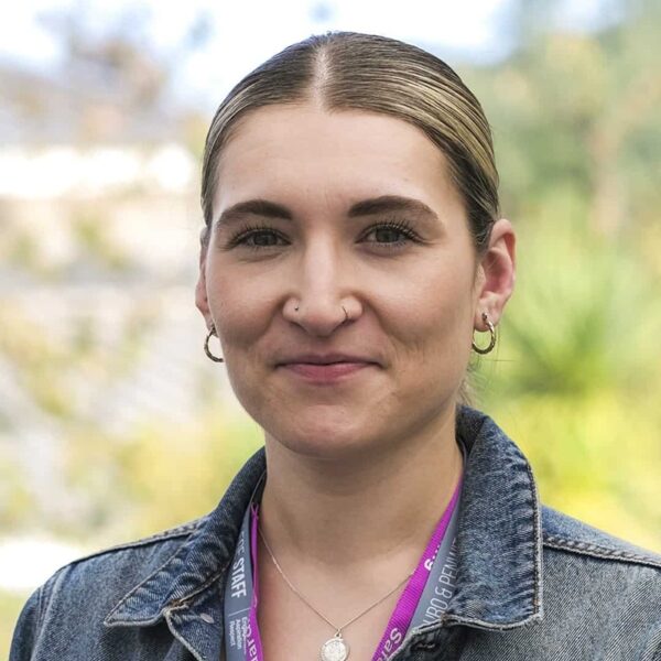 A photo of Jodie stood outside. She is wearing a denim jacket and two staff lanyards.