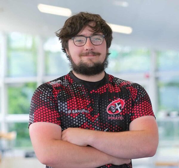 A young male student stands with his arms folded across his chest. He is wearing a black and red cornish pirates rugby top.