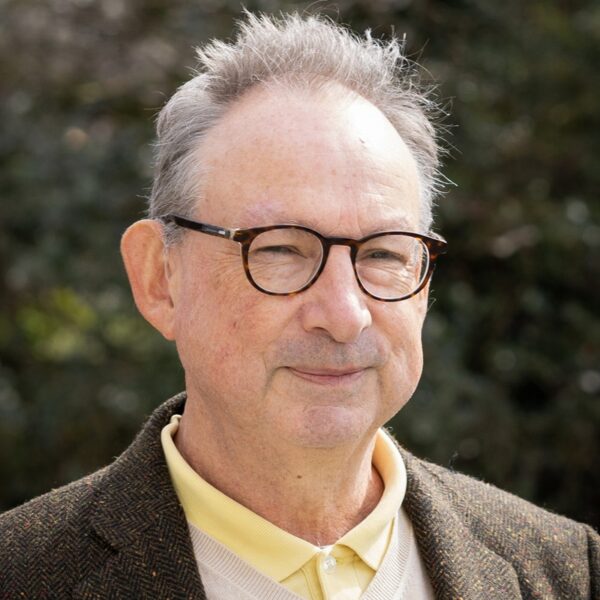 A portrait photo of Ken Curtis. He is wearing a yellow shirt, beige jumper and brown jacket.