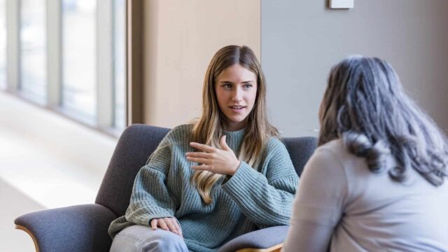 A young woman sits opposite an unrecognisable person on a comfy chair.