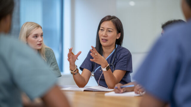 A small group of Nurses sit at a boardroom table as they meet to discuss patient cases. They are each dressed professionally in scrubs and have stethoscopes around their necks as they discuss plans of care for each case.