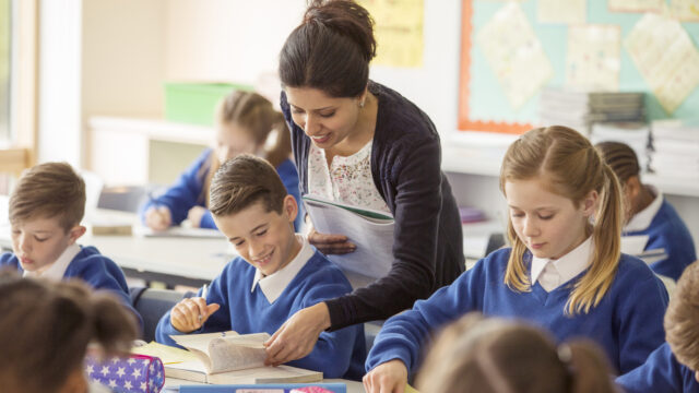 A teacher with pupils in a classroom. They look through books and are wearing a blue school uniform.