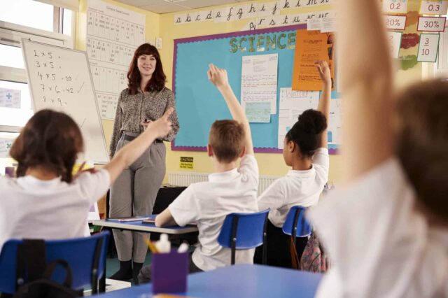 A classroom of students sit opposite a teacher with their hands held up ready to answer a question.
