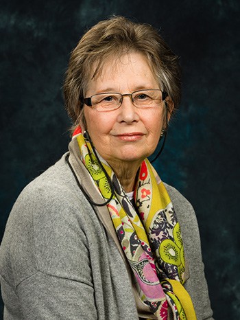 A photo of Linda Batchelor, she is wearing a grey cardigan, a colourful scarf and is sat against a dark backdrop.
