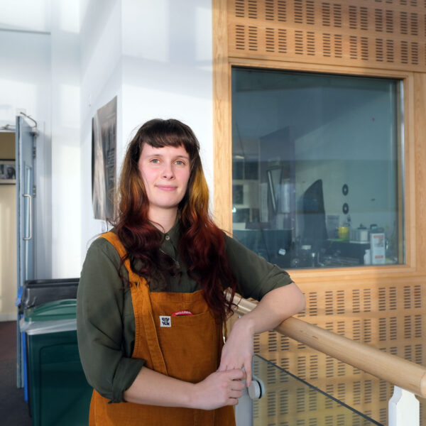 Lucy leans against a railing and is wearing orange dungarees.