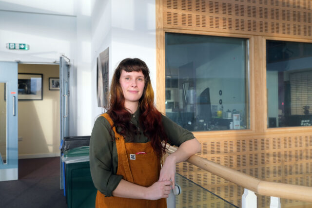 Lucy leans against a railing and is wearing orange dungarees.