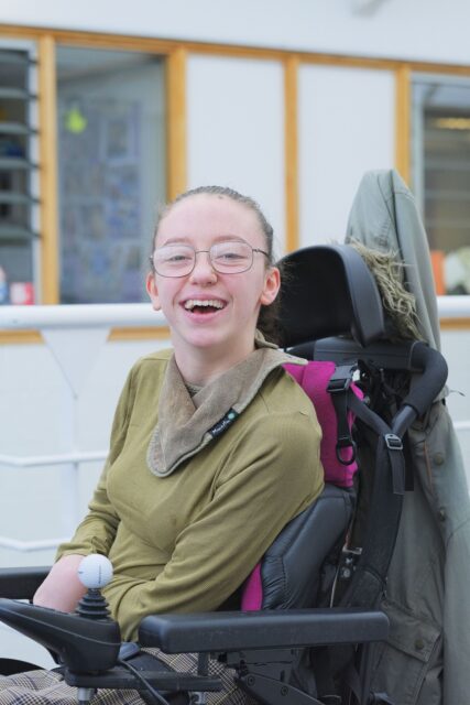 Maisie is sat in a wheelchair, wearing glasses and smiling.