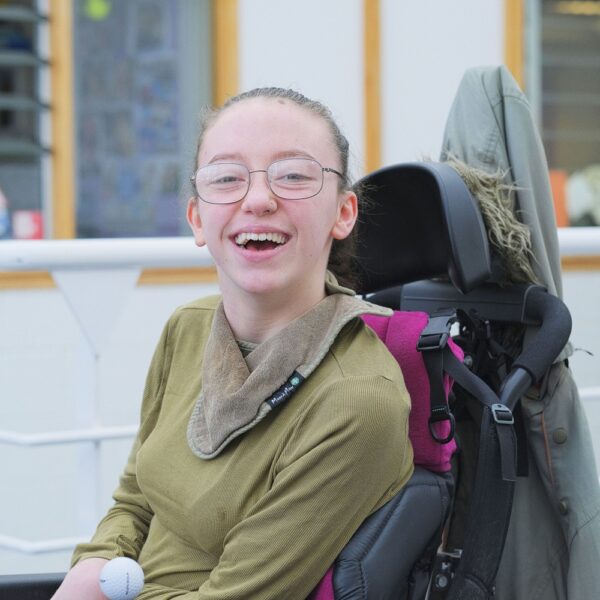 Maisie is sat in a wheelchair, wearing glasses and smiling.
