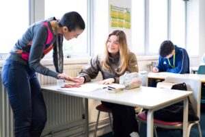 A tutor stands opposite a student who is working at a classroom table with a notebook and pencil case.
