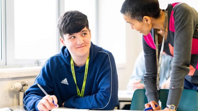 A tutor stands next to a student sat at a table with pen and paper.