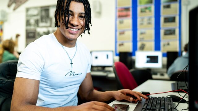 A student sat at a computer. He's turned and smiling at the camera.