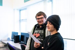 Two students look at a piece of paper in a computer room.