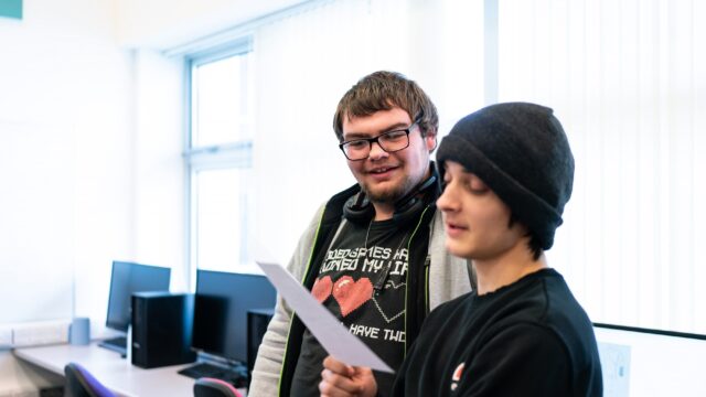 Two students look at a piece of paper in a computer room.