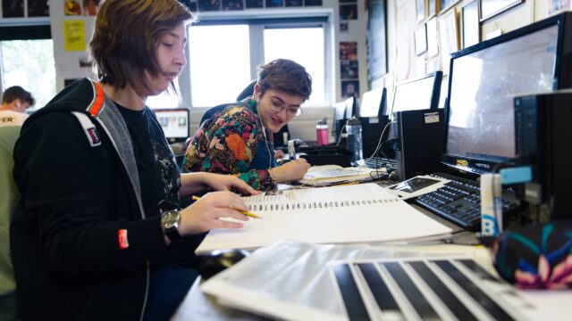 Students working at computers with large notebooks open in front of them.