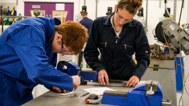 Students in overalls make measurements with tools and equipment surrounding them.