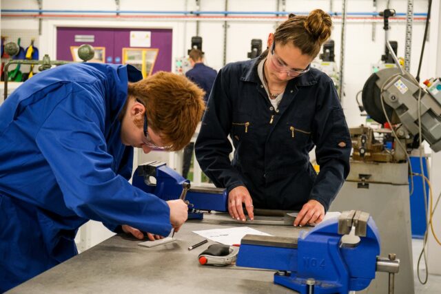 Students in overalls make measurements with tools and equipment surrounding them.