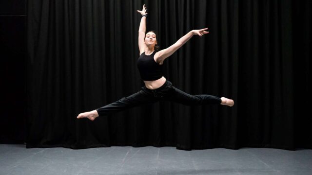A student leaping into the air with her arms and legs extended in a dance studio.