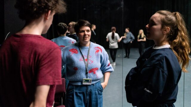 A tutor standing with students in a drama studio with large mirrors.