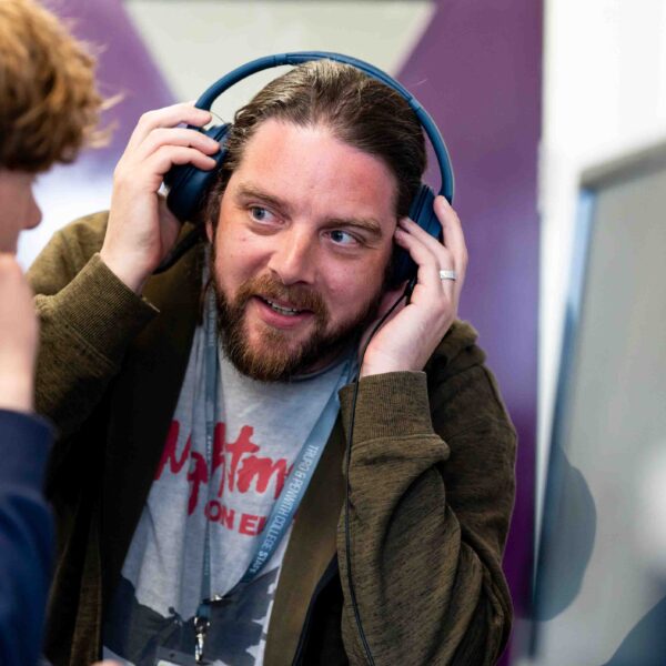 A tutor looks to a student and holds a set of headphones to his ears. The student is sat in front of a computer.