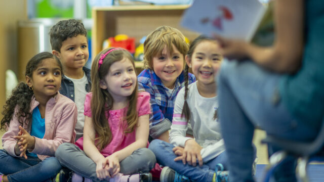 Young children sit on the floor cross-legged with a teacher in front of them.