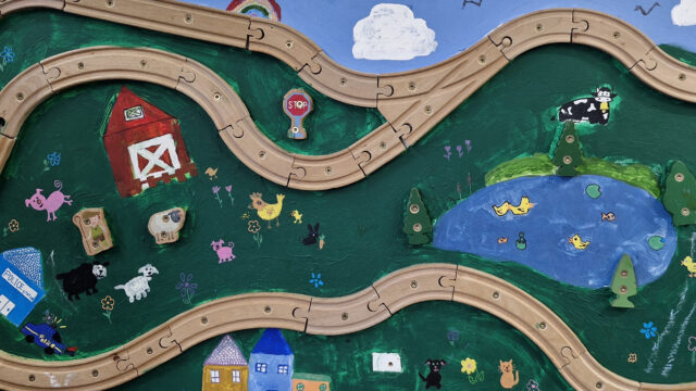 A board has been painted with fields, flowers, animals, a pond and houses. There is a wooden track going around the board.