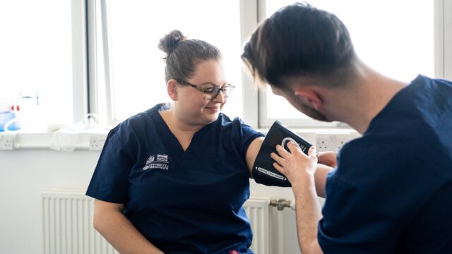 Two students in scrubs use a blood pressure monitor.