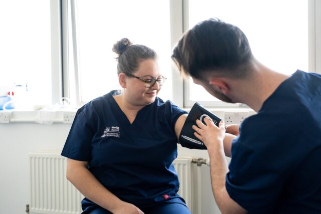Two students in scrubs use a blood pressure monitor.