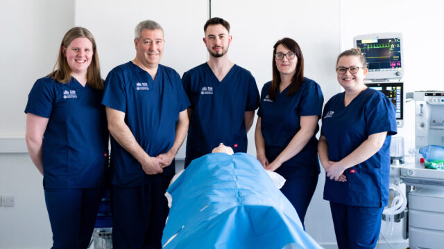 A group of medical students stand in a row in front of a dummy on an operating table.