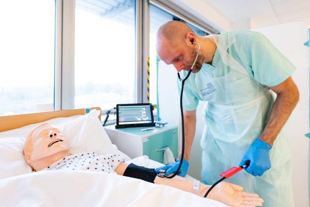 A student nurse holds a stethoscope to a dummy's arm.