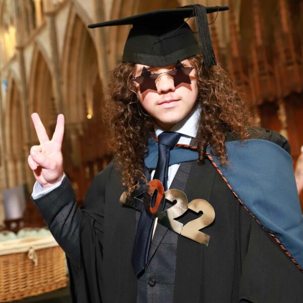 Ollie in graduation robes wearing star shaped sunglasses and holding his hand up in a peace sign.