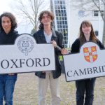 Three students stand outside holding two boards. One for Oxford and the other for Cambridge. The logos are on the boards.