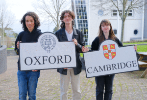 Three students stand outside holding two boards. One for Oxford and the other for Cambridge. The logos are on the boards.