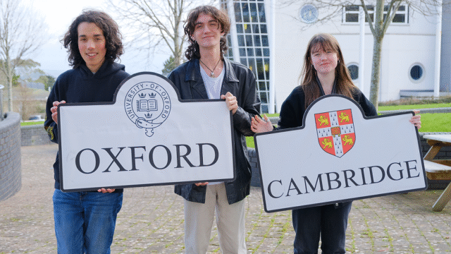 Three students stand outside holding two boards. One for Oxford and the other for Cambridge. The logos are on the boards.