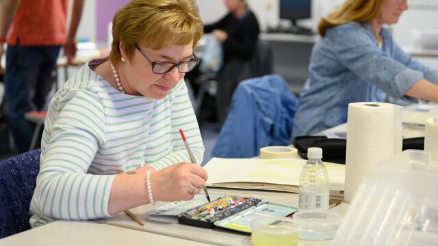 An adult student is painting with watercolours at a classroom table.
