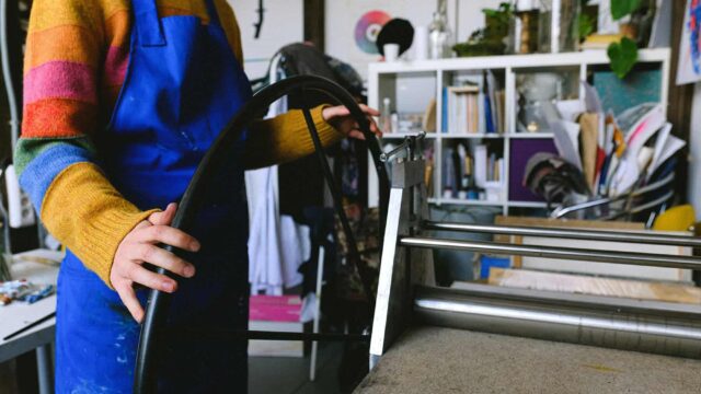 A close up of an art student wearing overalls using a wheel on a printmaking machine.