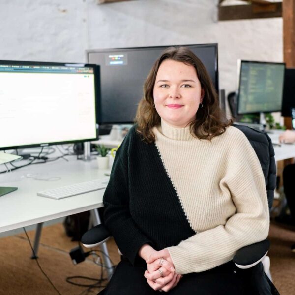 Paige is sat in an office with a long desk behind her and other colleagues working in the background.