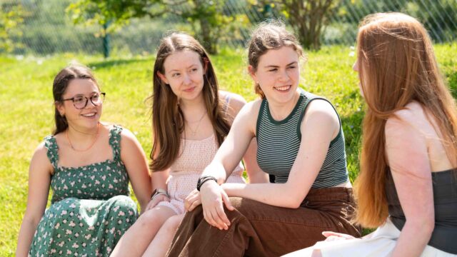 Four students sit on the grass outside and chat. It's a sunny day and they are all in dresses or t-shirts and jeans.