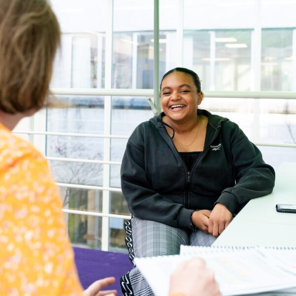 A student sits across from a member of staff. The student looks over the shoulder of the staff member towards the camera and smiles.