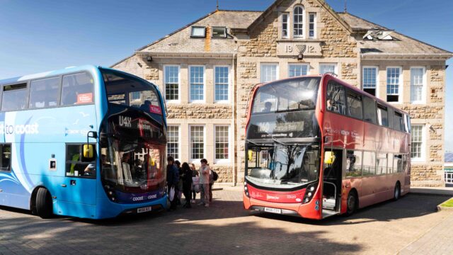 Two buses are parked in front of a college building and students are hopping onboard.