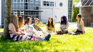 A group of students sit on the grass outside a college building. They have daisies surrounding them and one student has daisies in her hair.