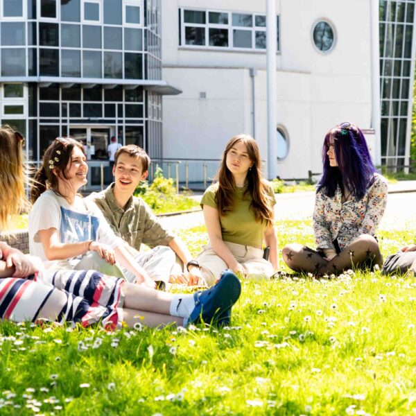 A group of students sit on the grass outside a college building. They have daisies surrounding them and one student has daisies in her hair.