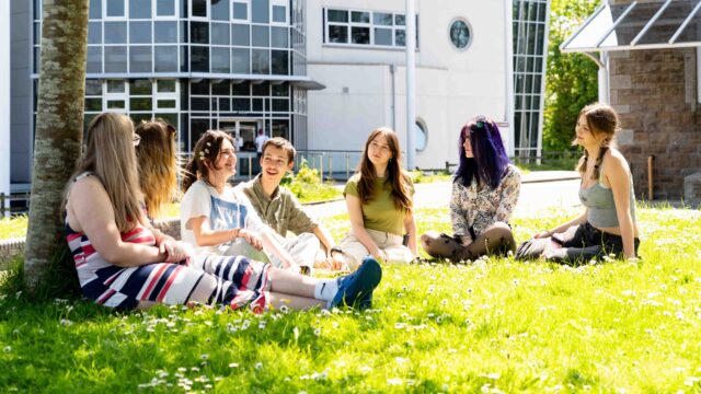 A group of students sit on the grass outside a college building. They have daisies surrounding them and one student has daisies in her hair.
