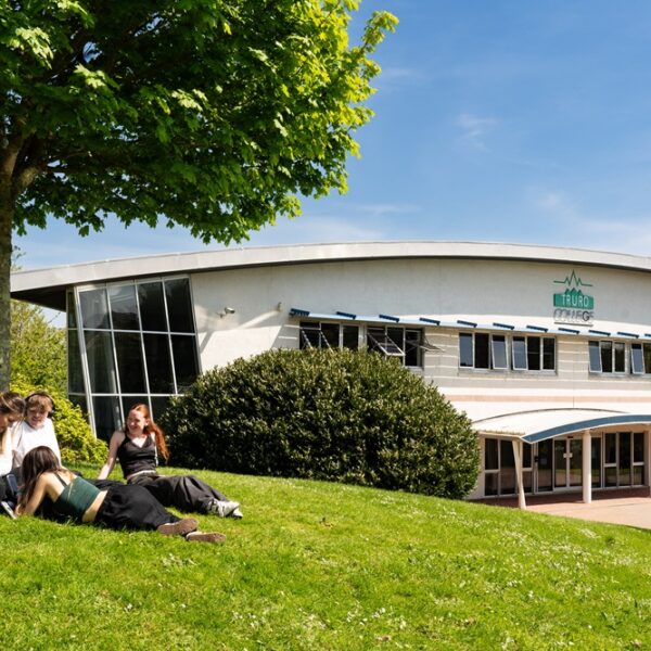 Four students sit out on the grass under a tree at Truro College. It is a sunny day and the Helford building is in the background.