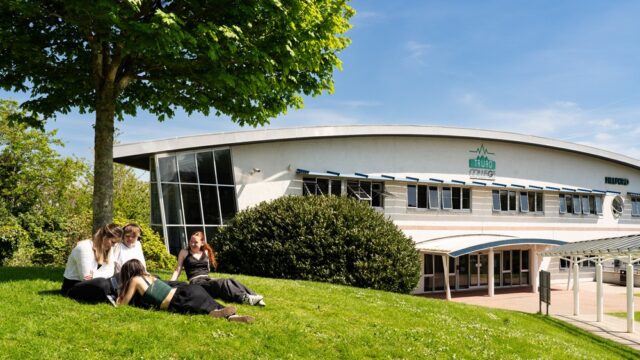 Four students sit out on the grass under a tree at Truro College. It is a sunny day and the Helford building is in the background.