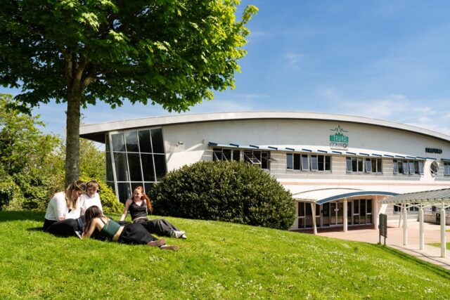 Four students sit out on the grass under a tree at Truro College. It is a sunny day and the Helford building is in the background.