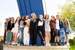 A group of students line up and stand on a low wall outside. They have their arms around each other.