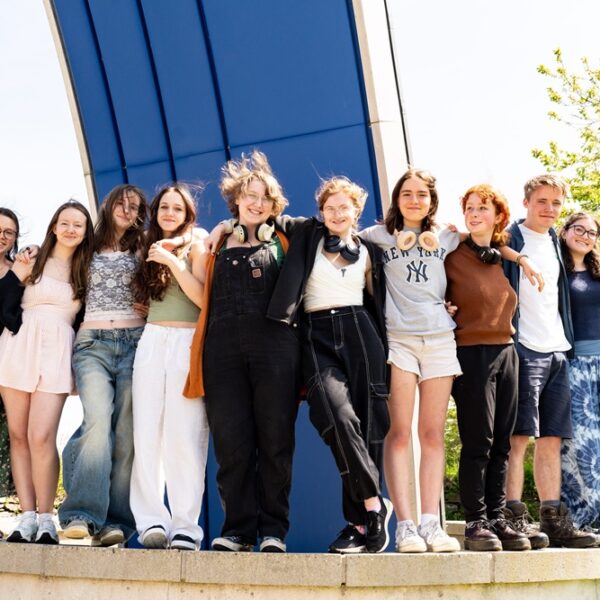 A group of students line up and stand on a low wall outside. They have their arms around each other.