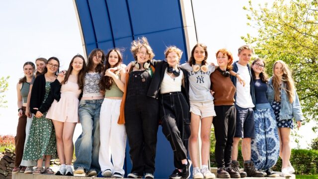 A group of students line up and stand on a low wall outside. They have their arms around each other.